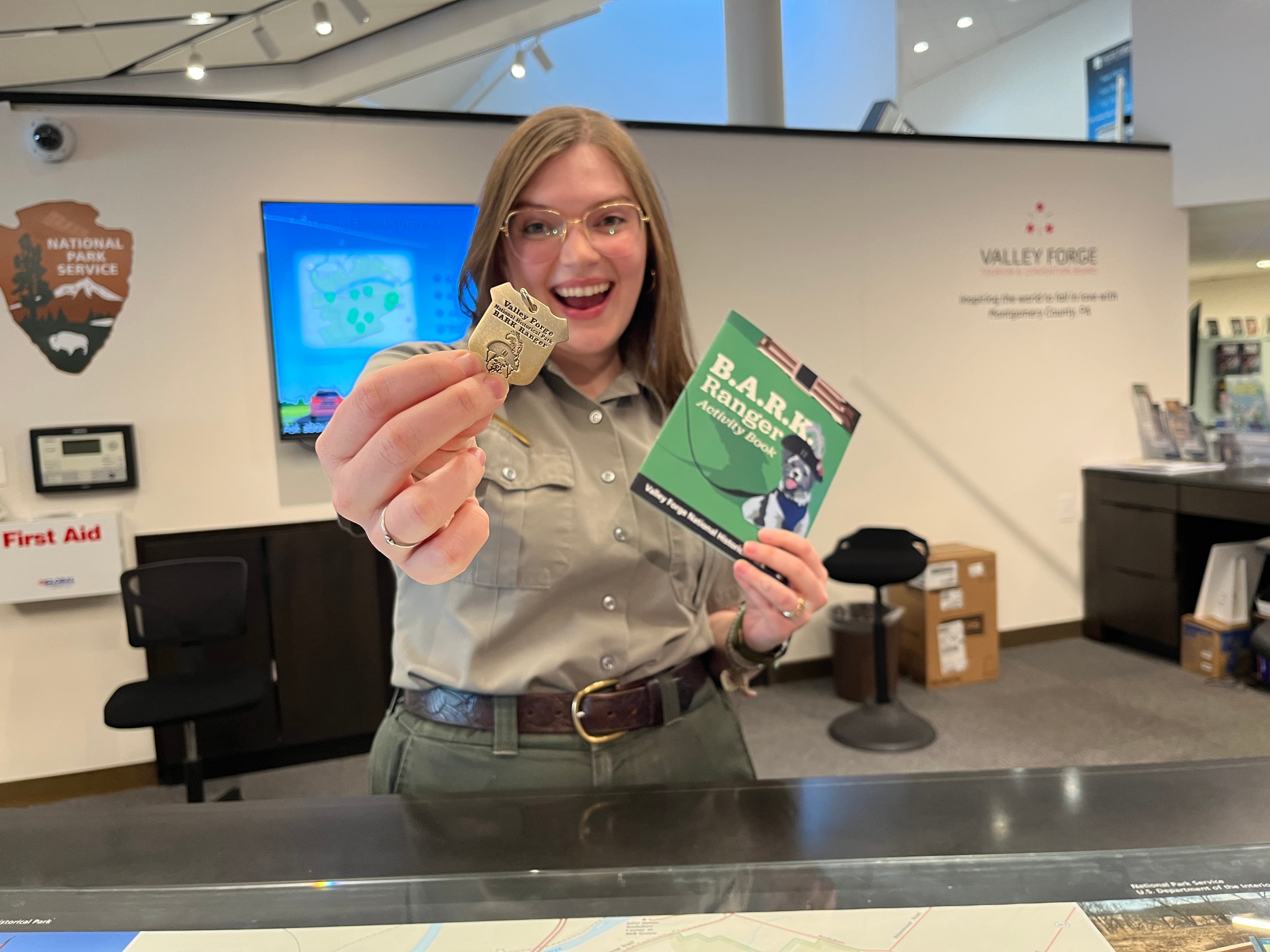 A smiling park ranger holds out a B.A.R.K. Ranger dog tag and activity book
