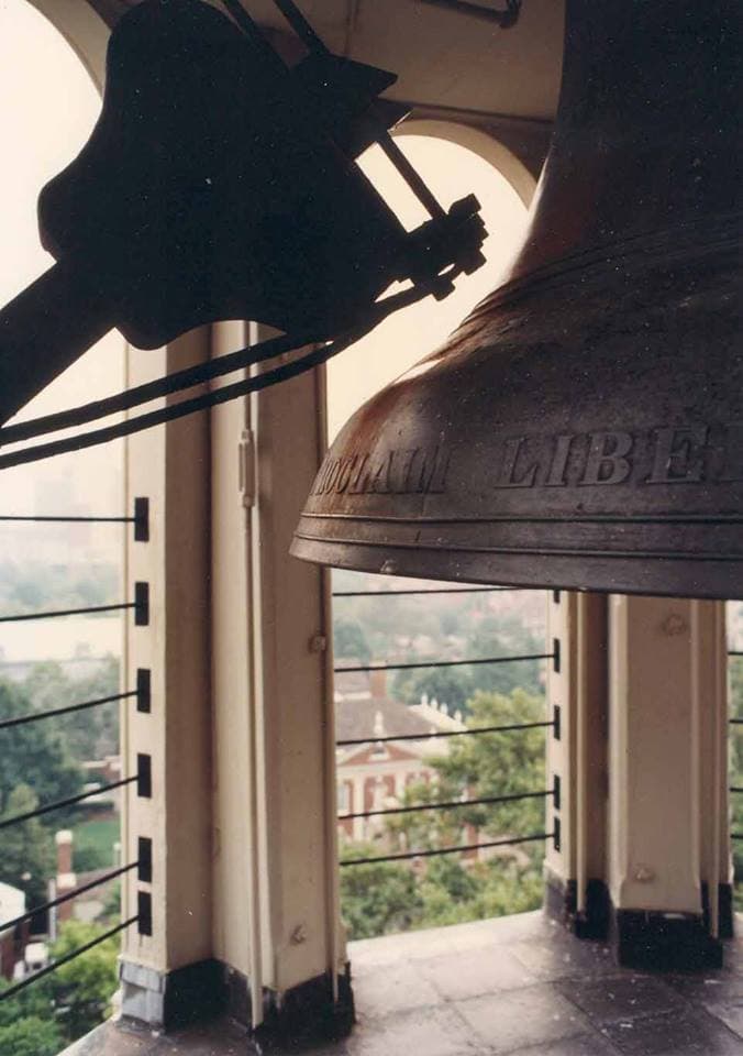 A hammer mechanism sits to the side of the base of a large bell in a tower overlooking city building