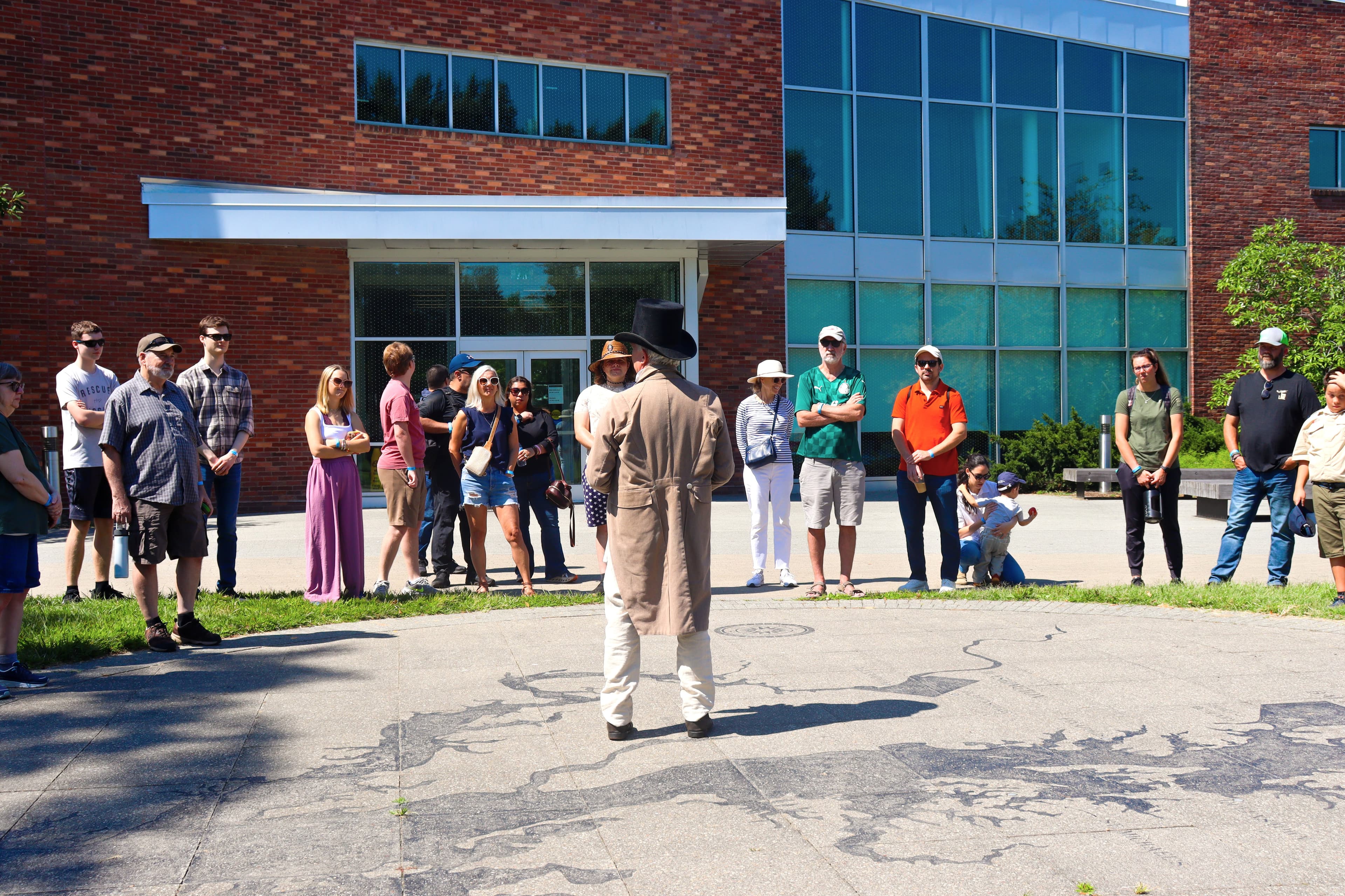 Ranger in living history garb speaks to a semicircle of people outside of the visitor center.