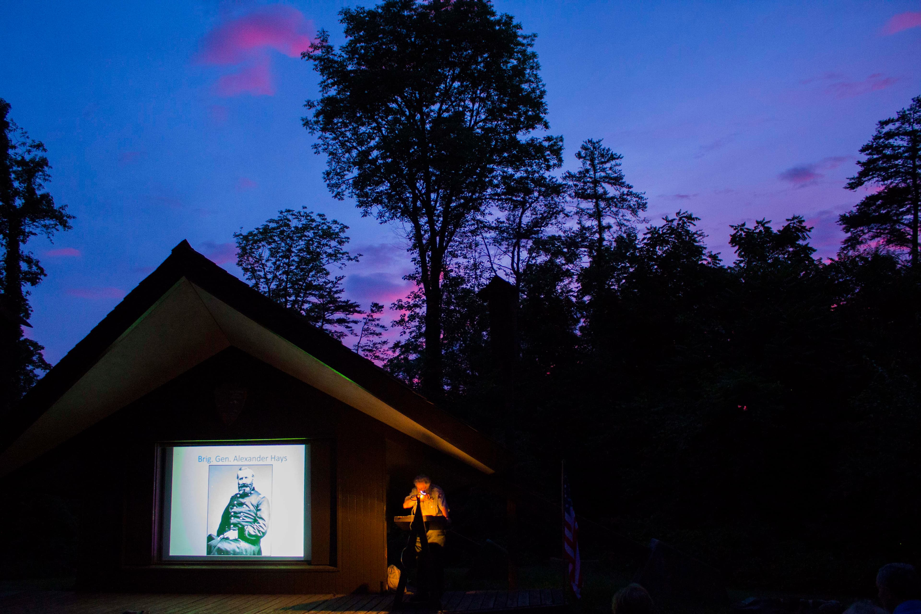 A park ranger at an amphitheater speaking with a slide show next to him.