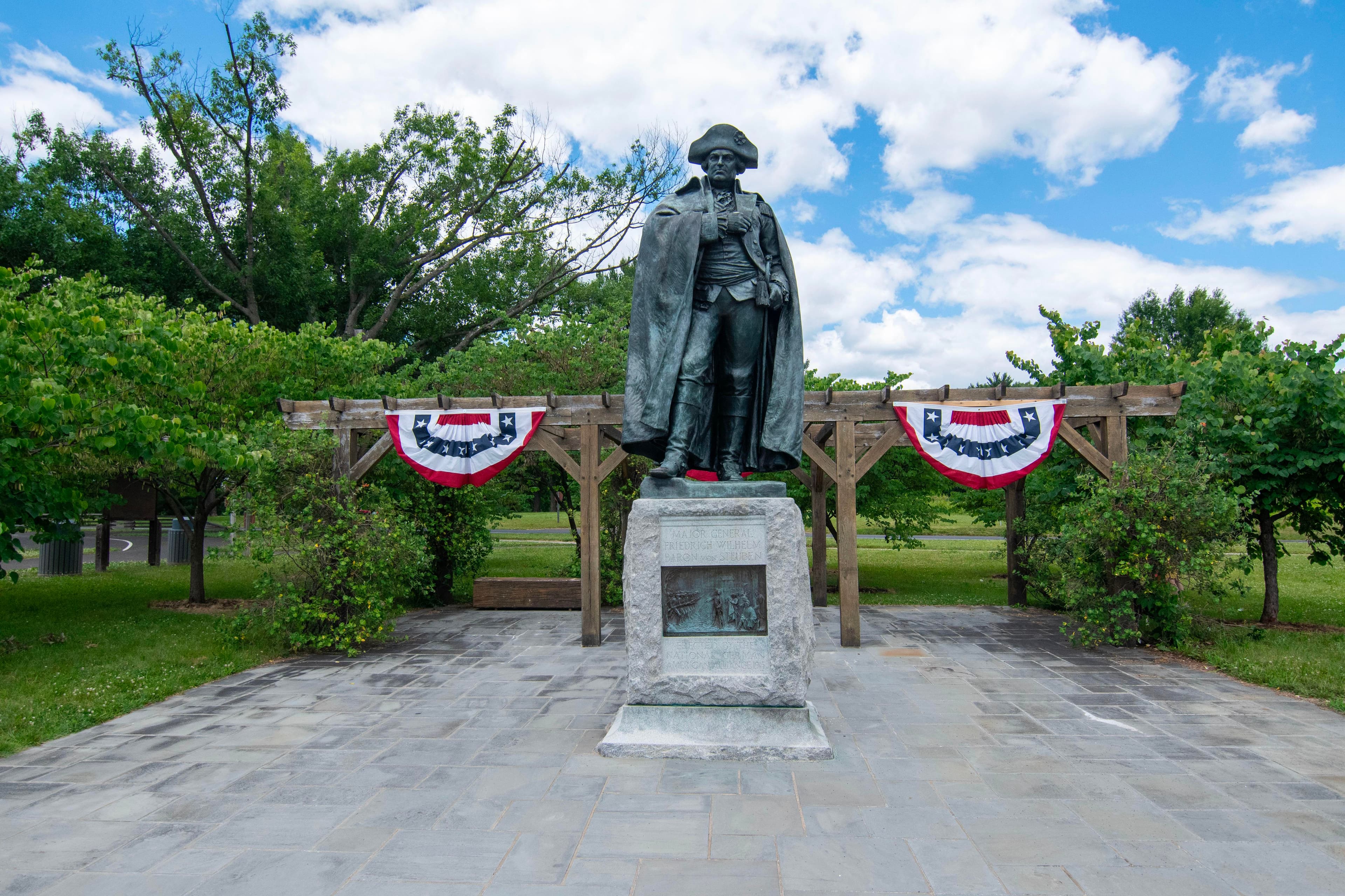 bronze statue of rev war general on stone base, red white and blue bunting behind